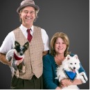 An older man and woman pose together in front of a neutral the woman is holding a small white dog