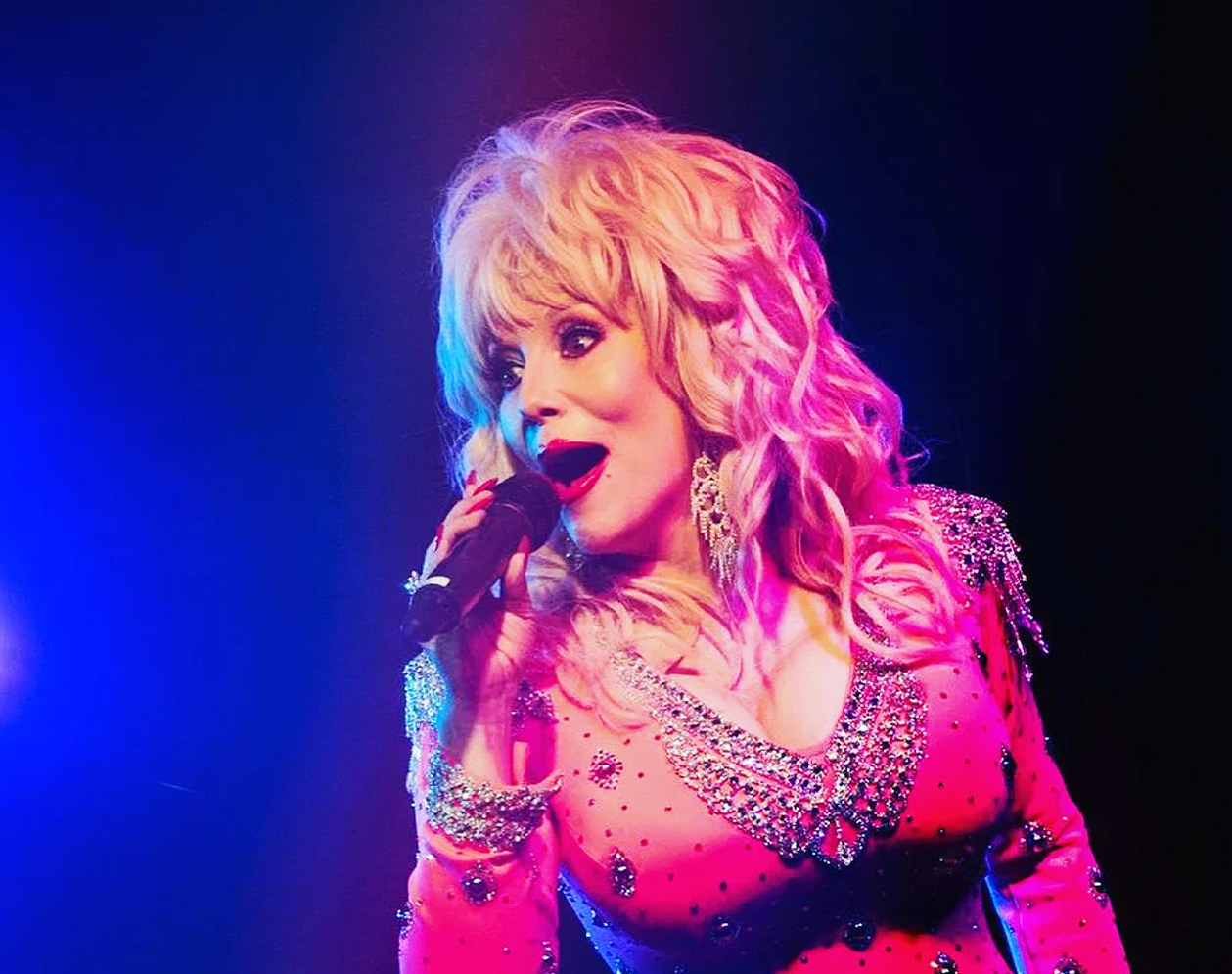 A woman holding a mic and singing. She is wearing a Dolly Parton style wig, makeup and rhinestone studded hot pink dress. 