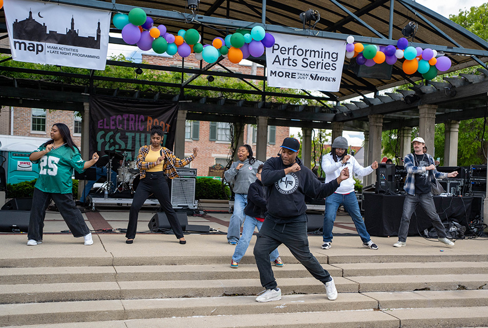 A black man stands on the steps of the Oxford Uptown Park Pavillion teaching hip hop dance moves to a crowd of people standing in front of him of a green lawn.