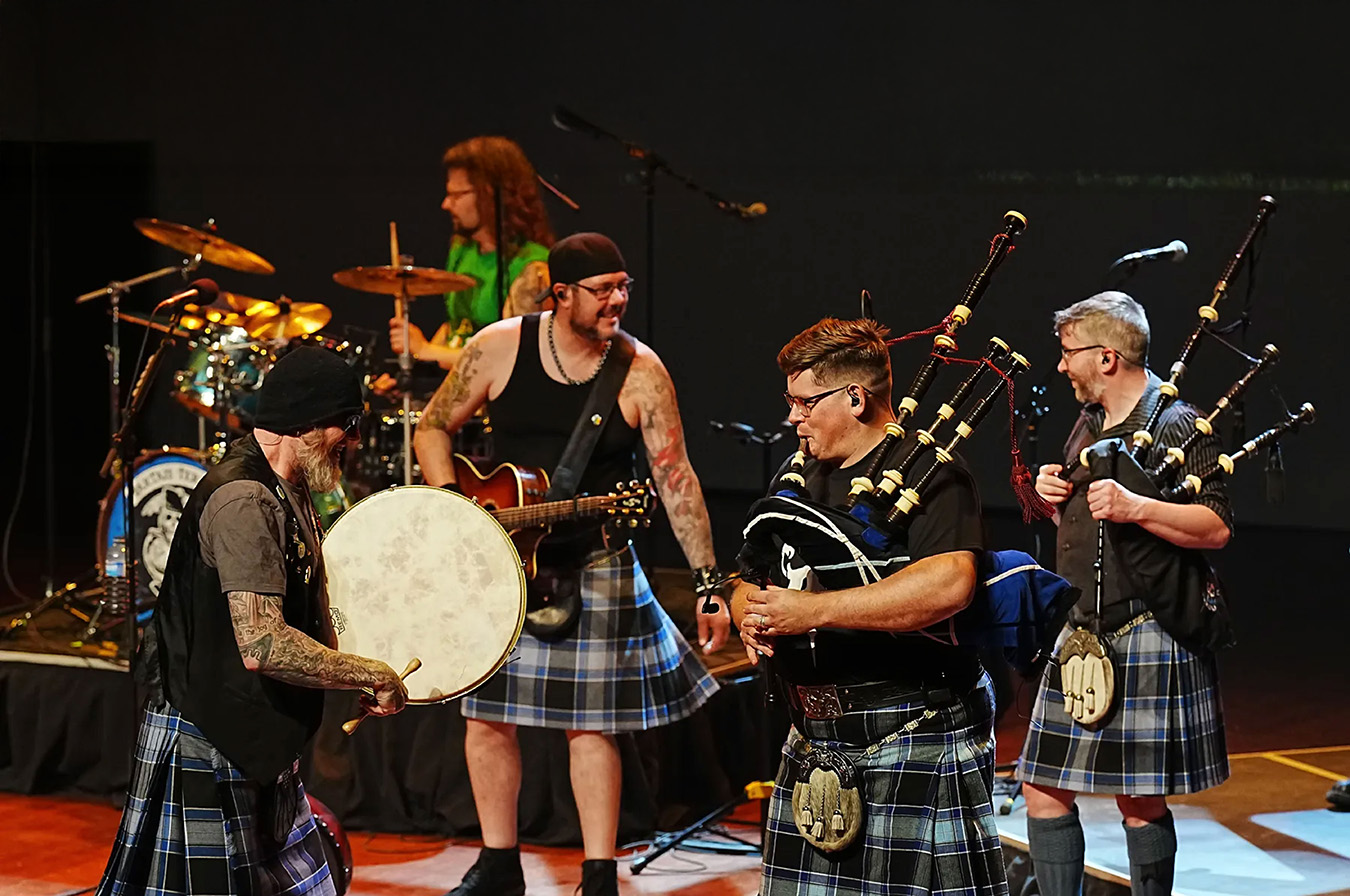 A photo of a band performing live on stage. The band members are all wearing kilts and kilt hose. One is playing guitar. One is playing a rock style drum kit. Two are playing bagpipes and one is playing a bodhran drum.