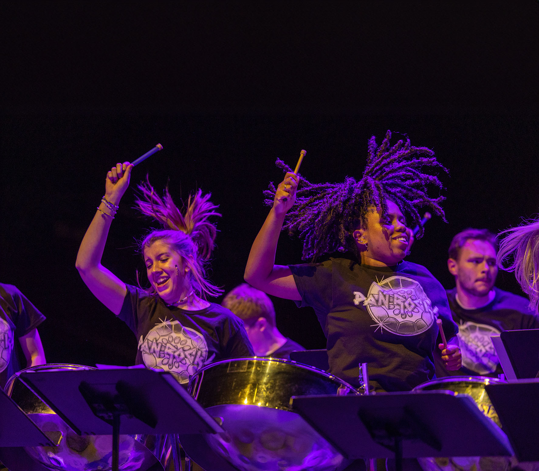 A photo of three college age women mid jump each standing behind a steel pan drum with more players behind them and in a dark room with purple and blue rock concert lighting effects