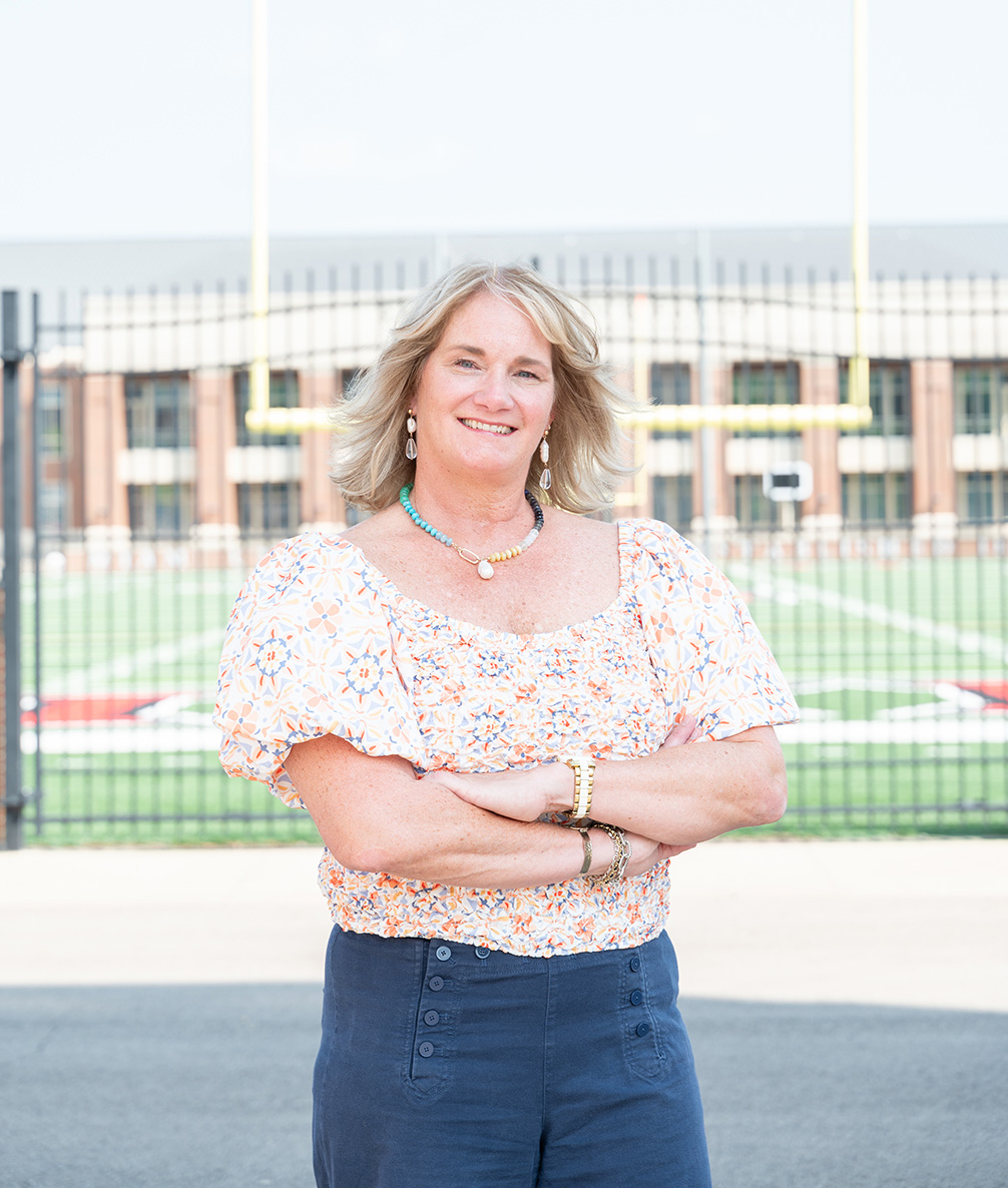 Photo of Missy Friede, a woman with curly blnd hair wearing a colorful peseant blouse and jeans standing at Yager Stadium