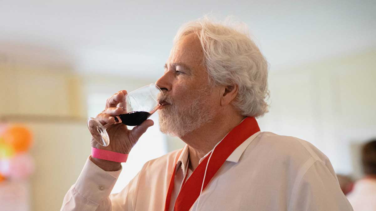 A man with wavy white hair and white beard brings a glass of red wine to his lips to taste