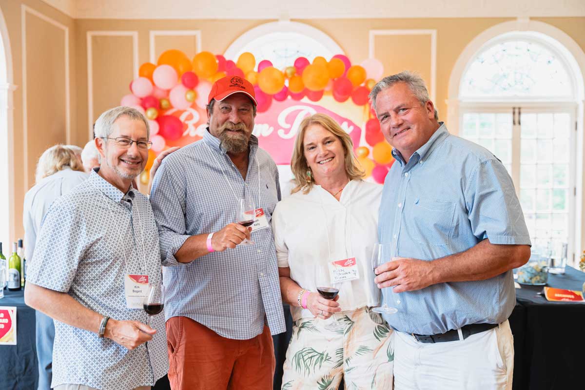 Four attendees holding wine glasses and smiling in front of a colorful display at the Oxford Community Arts Center.