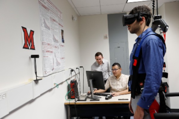 Male student is standing in the foreground, connected to a VR headset and other equipment. Dr. James Chagdes and another male student are in the background work on a computer.