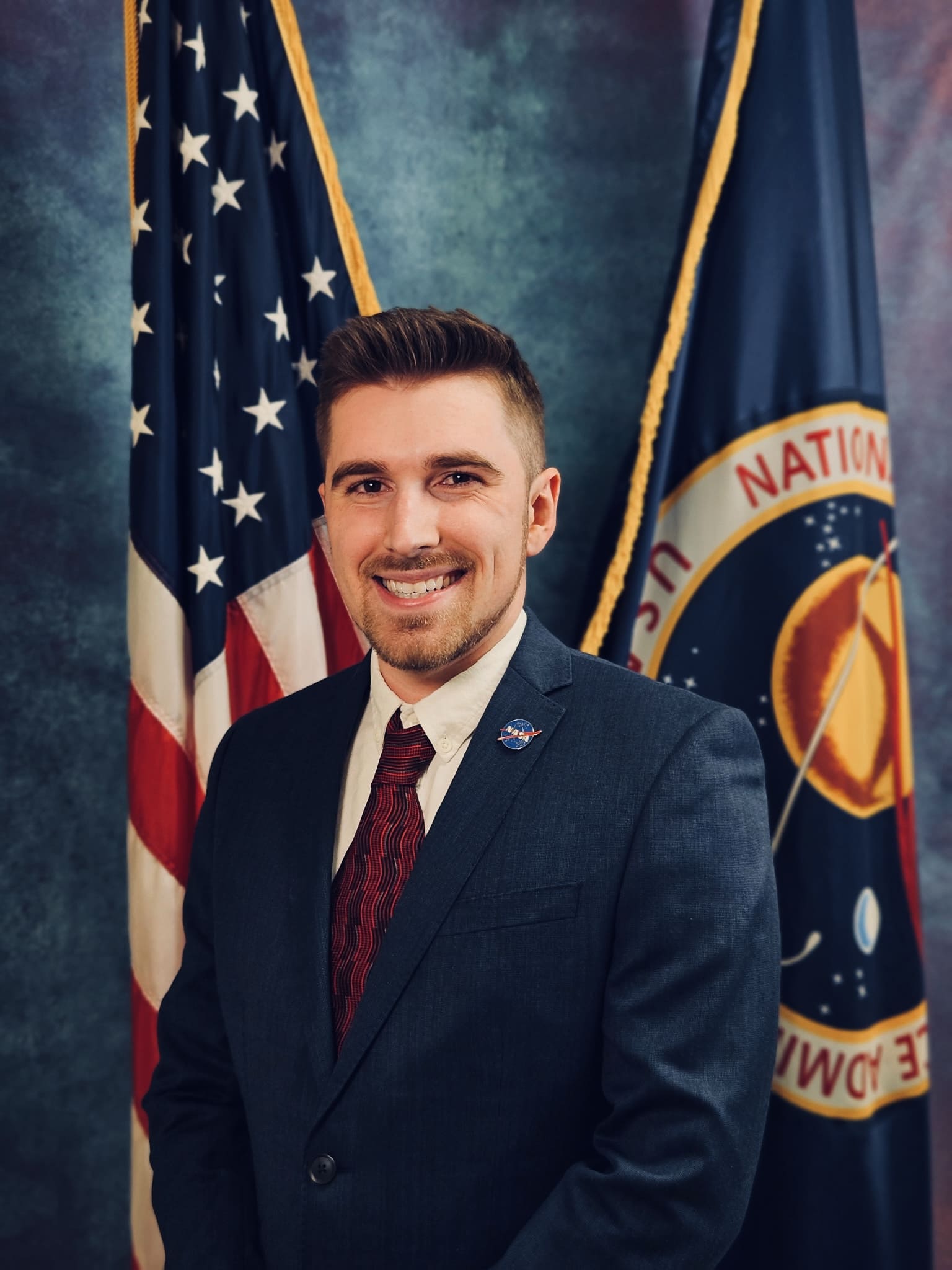 Smiling man in a suit with a NASA lapel pin, standing in front of the U.S. flag and a NASA flag.