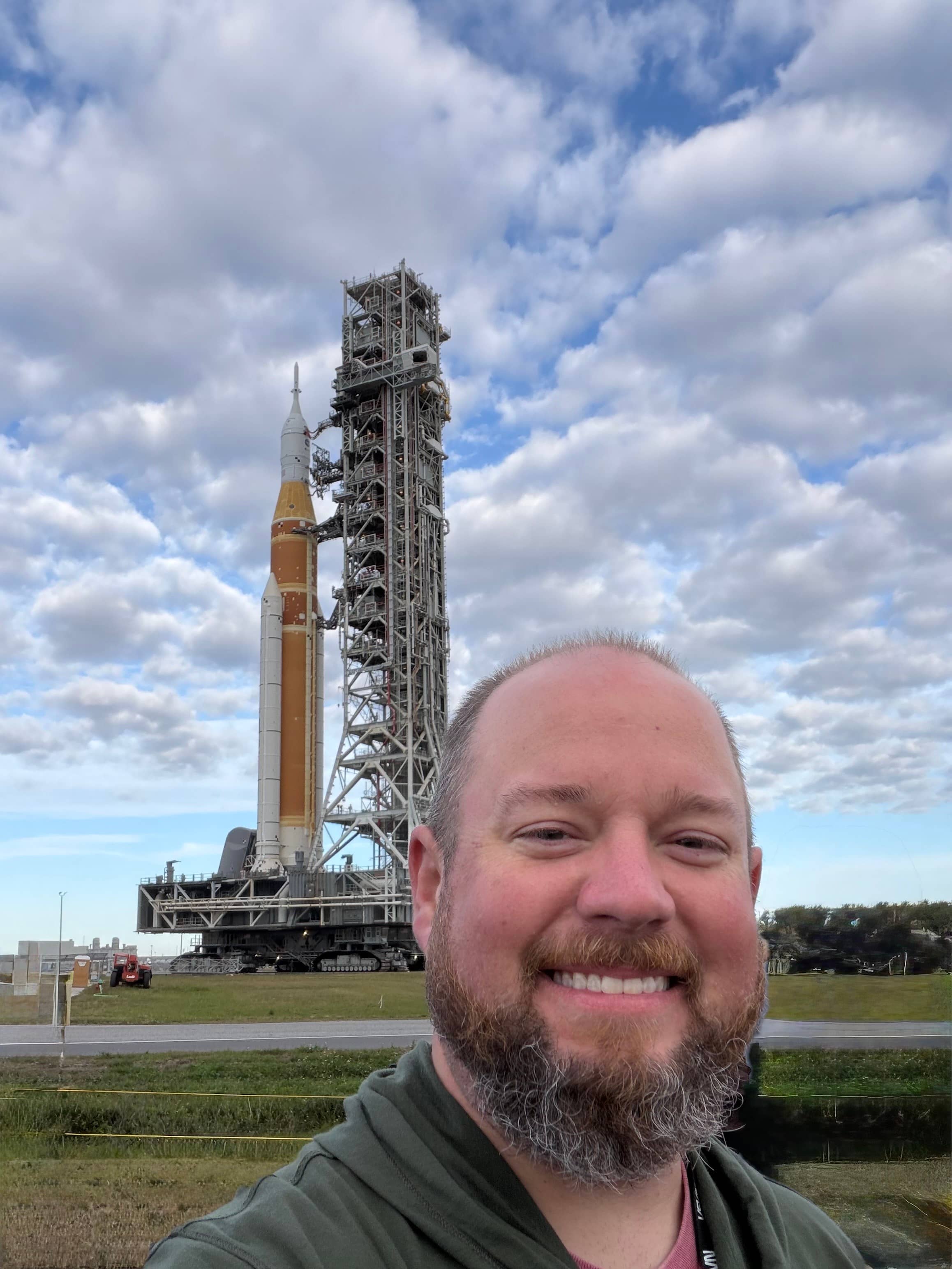 Smiling man taking a selfie in front of a rocket on its launch pad under a cloudy sky.