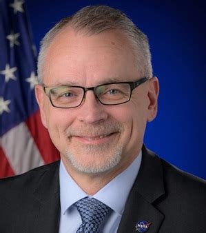 Smiling man in a suit and glasses in front of an American flag and a blue backdrop.