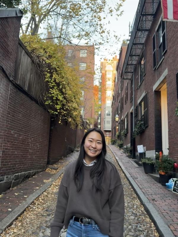 Smiling woman standing in a narrow cobblestone alley lined with brick buildings and autumn leaves.