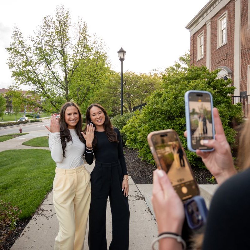 Two female students standing outside Benton Hall being photographed by another student.