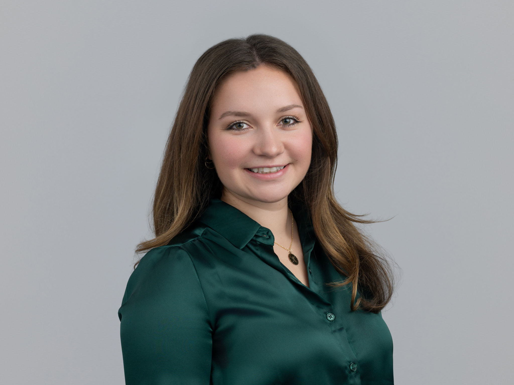 Smiling woman with long brown hair wearing a dark green blouse against a gray background.