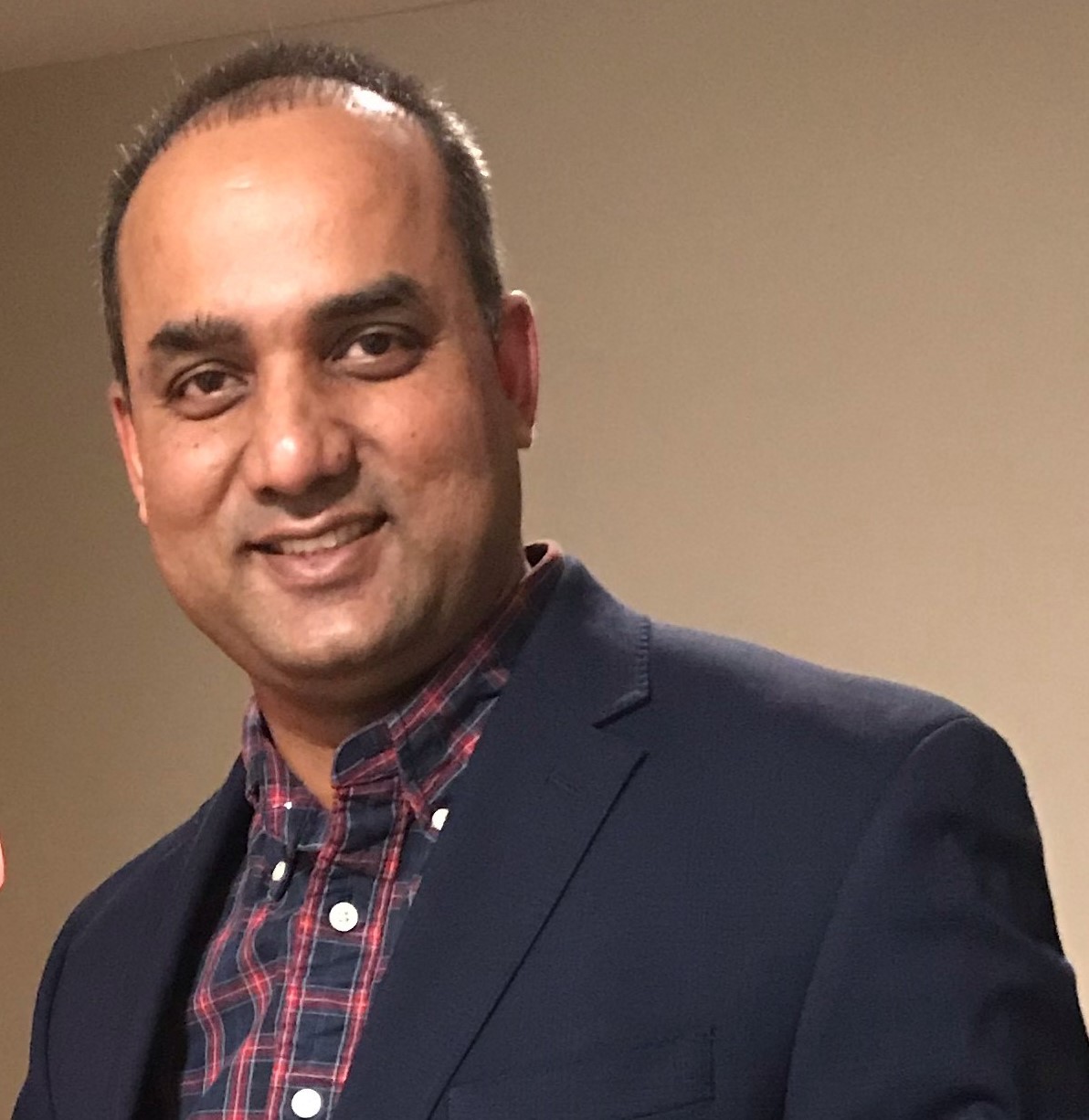 Smiling man in a navy blazer and plaid shirt against a plain indoor background.
