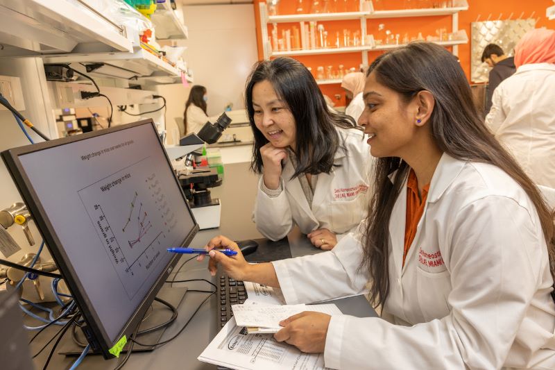 Two female students in lab coats looking at a graph on a computer