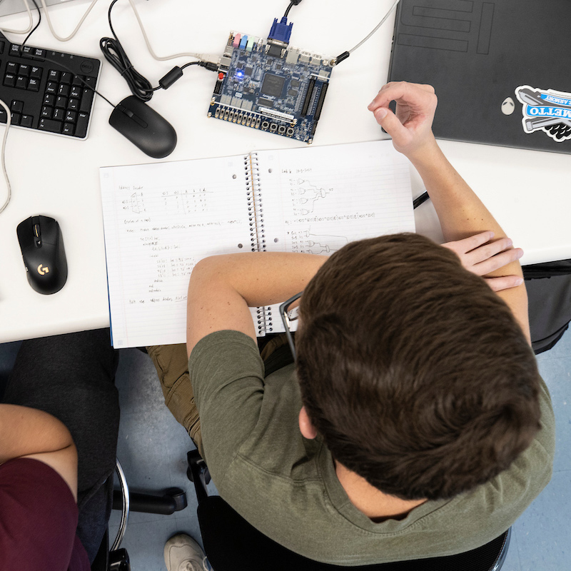 Overhead shot of two students working on laptops and a circuit board in a computer engineering class.