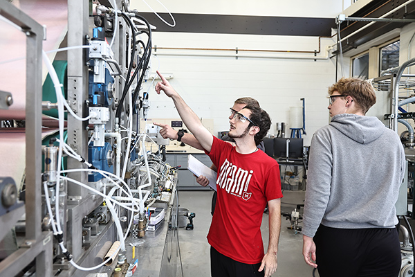 Two students indicate towards a piece of machinery in the pilot paper lab at Miami University while another walks by and takes notice.