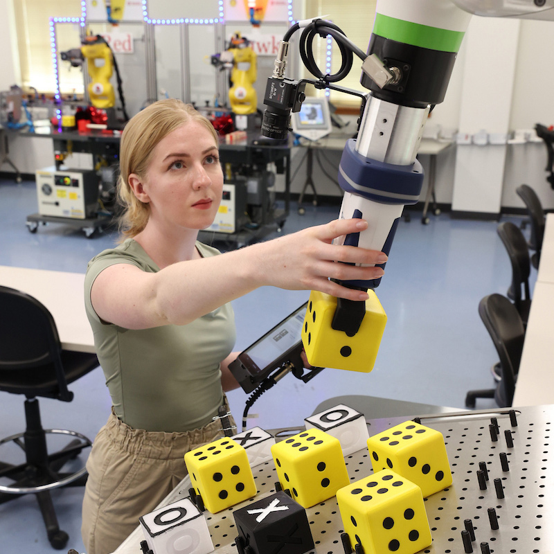 A female Robotics Engineering student works the controls of a FANUC industrial robot in the Miami University Industrial Robotics lab.