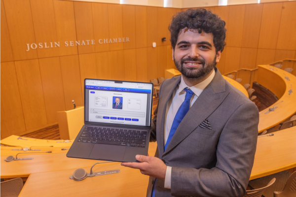 Sam Hieken stands in the Joslin Senate Chamber with a laptop displaying the Who Did I Elect? searchable database.