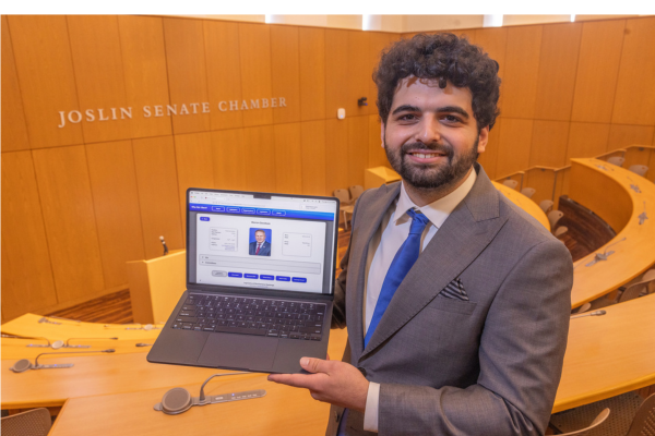 Sam Hieken stands in the Joslin Senate Chamber with a laptop displaying the "Who Did I Elect?" searchable database.