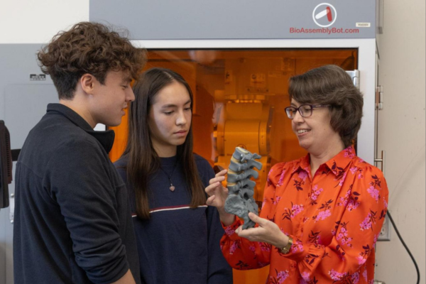 CPB Professor and Chair Jessica Sparks discusses a 3D spine model, noting the capabilities of the BioAssembly Bot 3D Printer located behind them, with Miami students in her research lab.