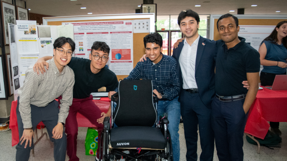 Five engineering students smile and pose around a uniquely engineered wheelchair.