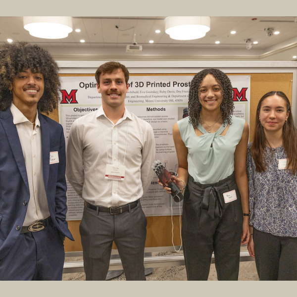 Team of undergraduate students stand in front of their research poster holding prosthetic hand