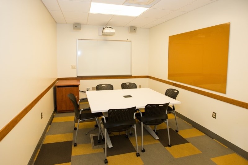Small room with a square table, 6 chairs around it. Two dry erase boards on the wall. 