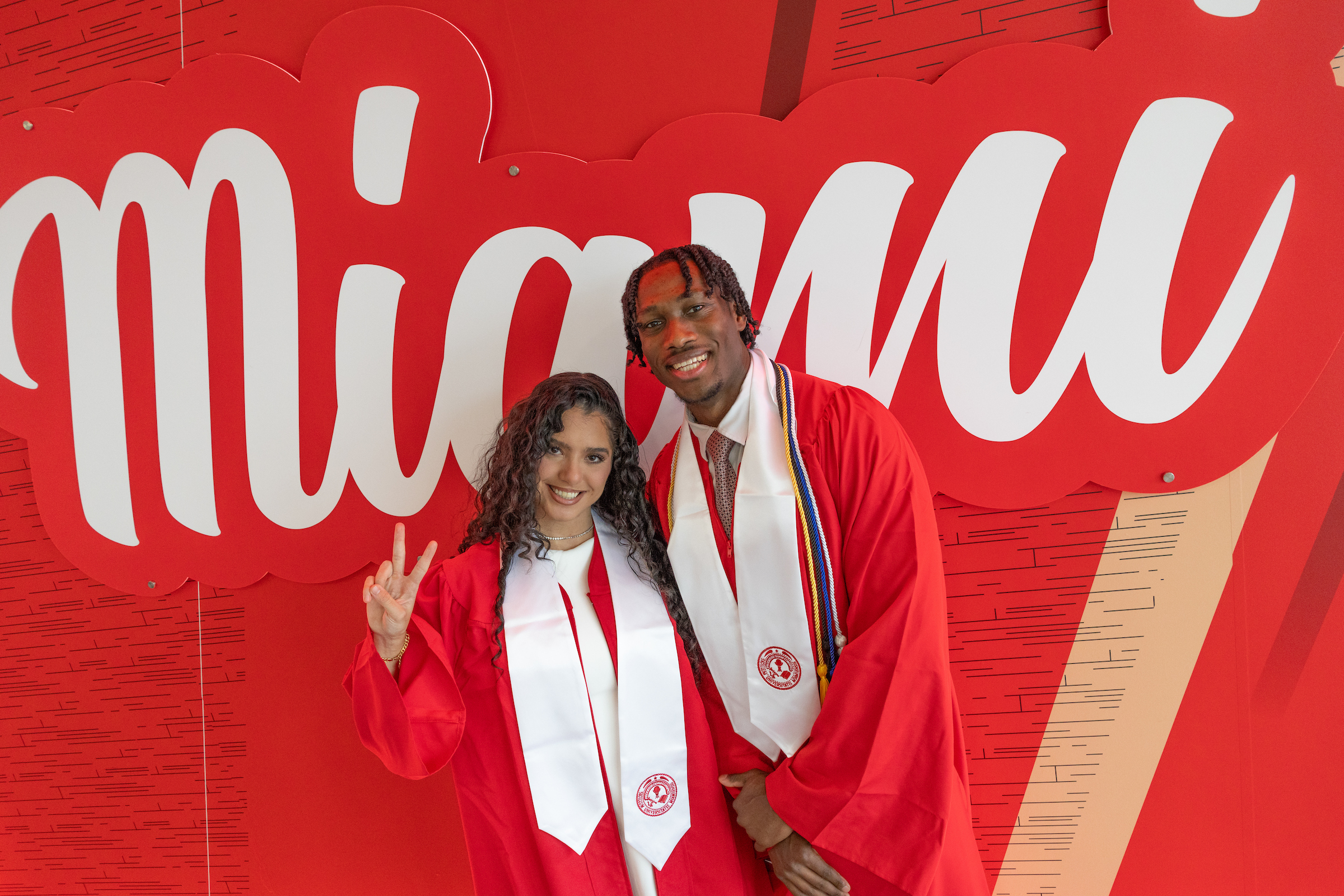 Two graduates pose in front of a wall graphic with the Miami script emblazoned on it. One gives a victory sign.