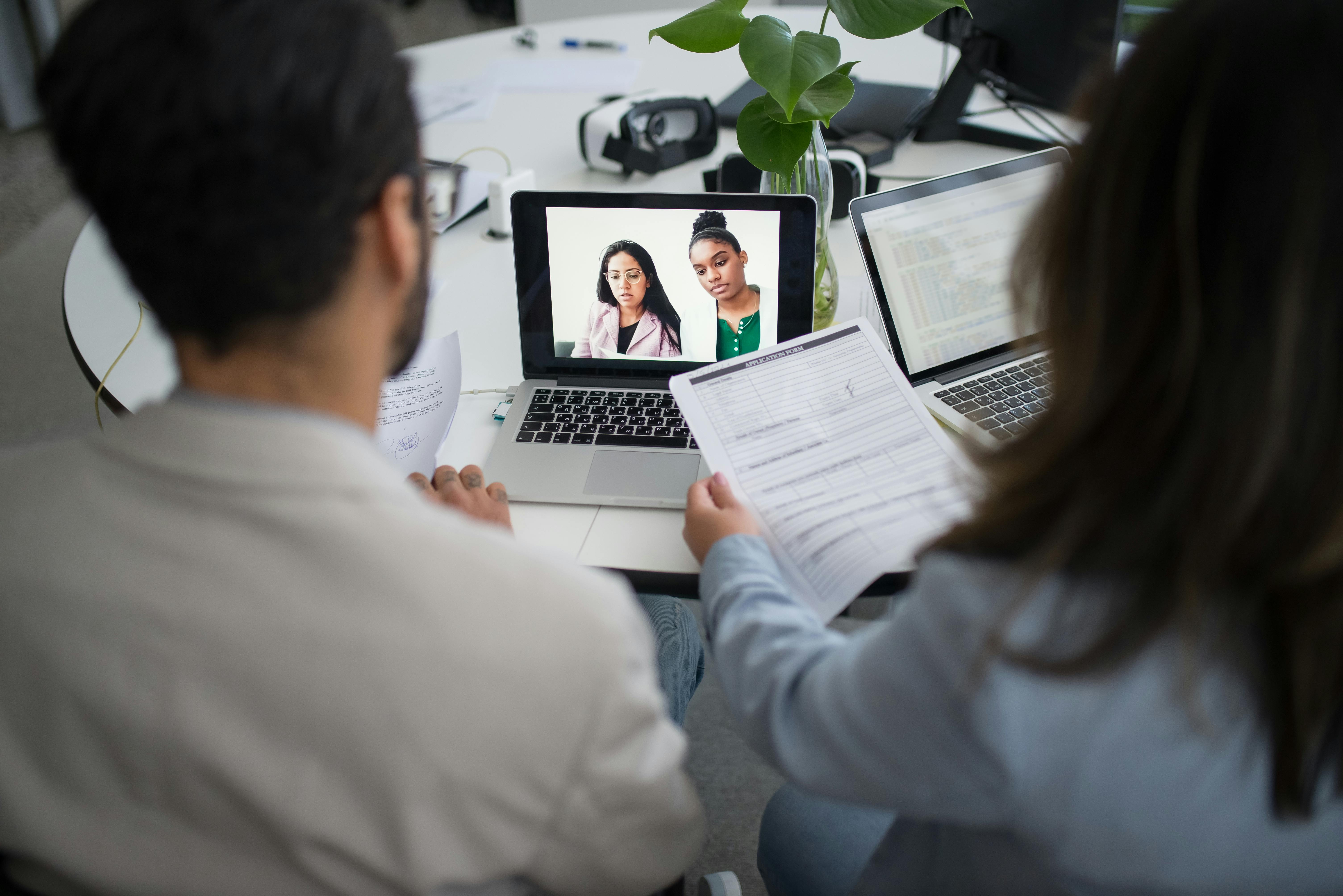 Four people engage in a virtual interview session, with two on a computer screen and two sitting at a desk.