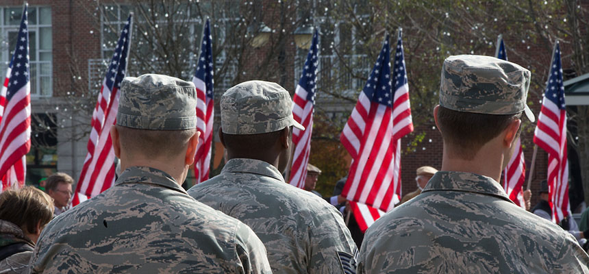 Three soldiers stand with their backs to the camera in front of American flags.