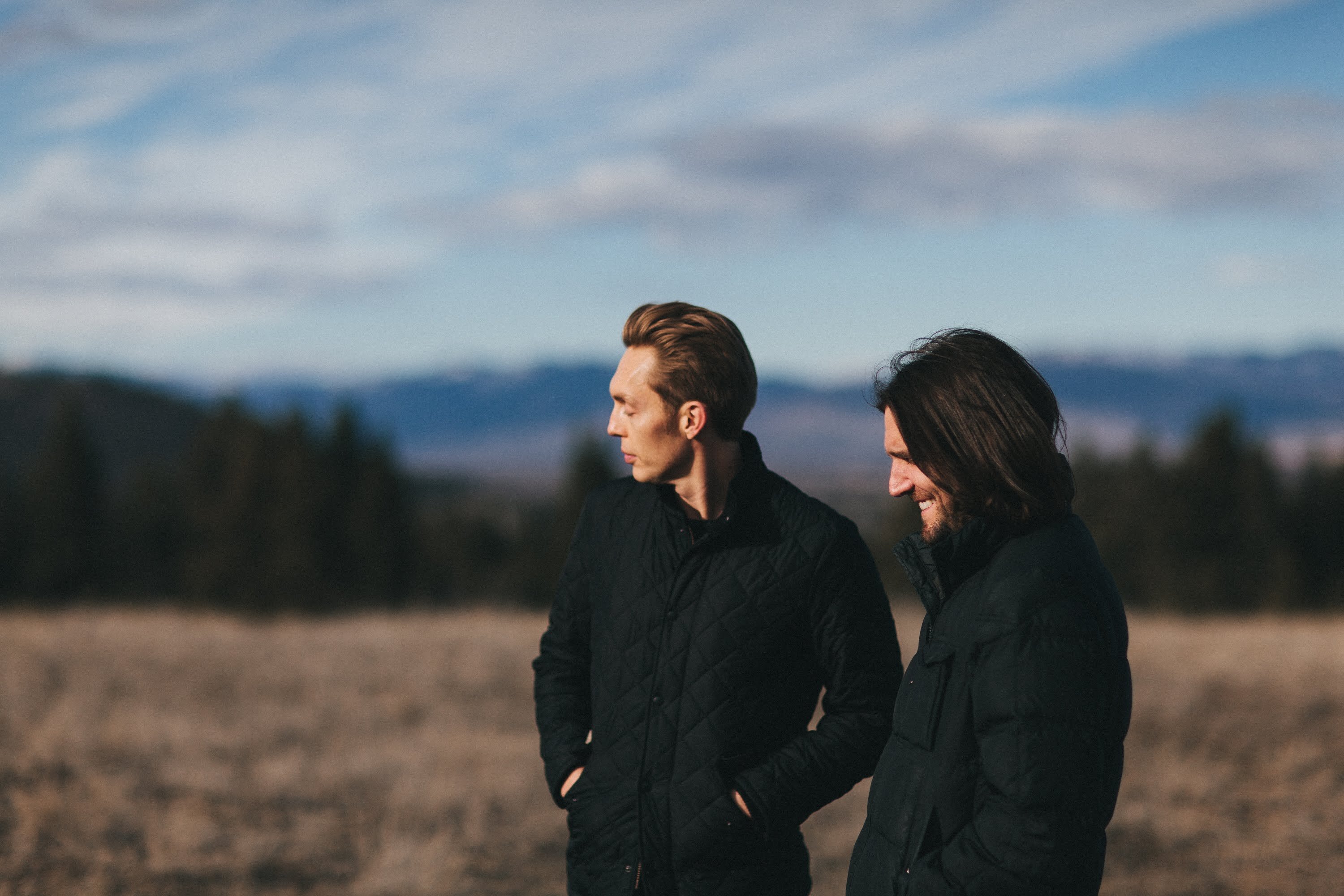 The Minimalists stand in a field in front of a clear blue sky.
