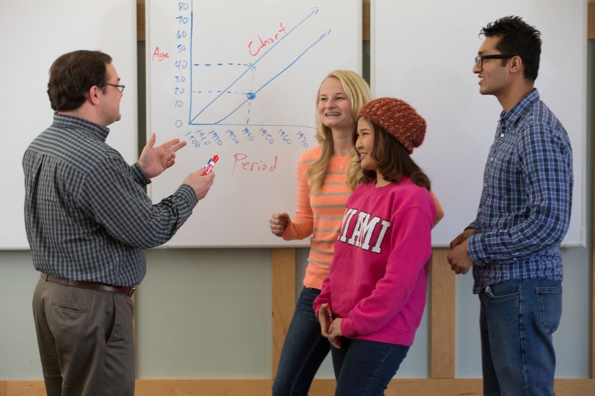 Professor talks with students in front of a whiteboard.