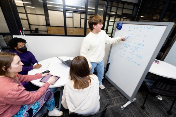 A student standing at a white board, three seated students paying attention.