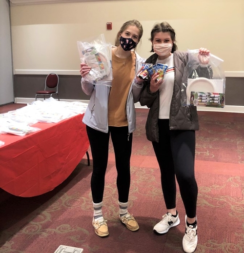 Two students in Armstrong Pavilion wearing masks. They are each holding up a bag of art supplies. A table of capris suns is on the table behind them. 