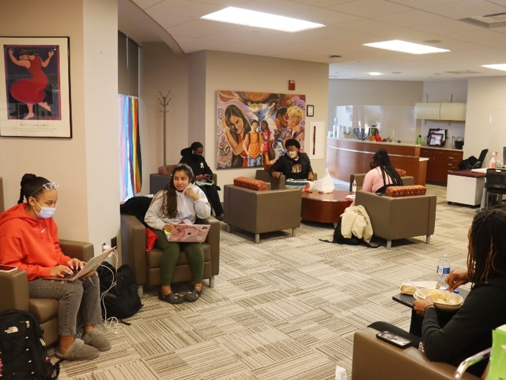 Center for Student Diversity and Inclusion. students in armchairs and laptops, eating lunch in the space. Vibrant paintings and posters are on the wall.