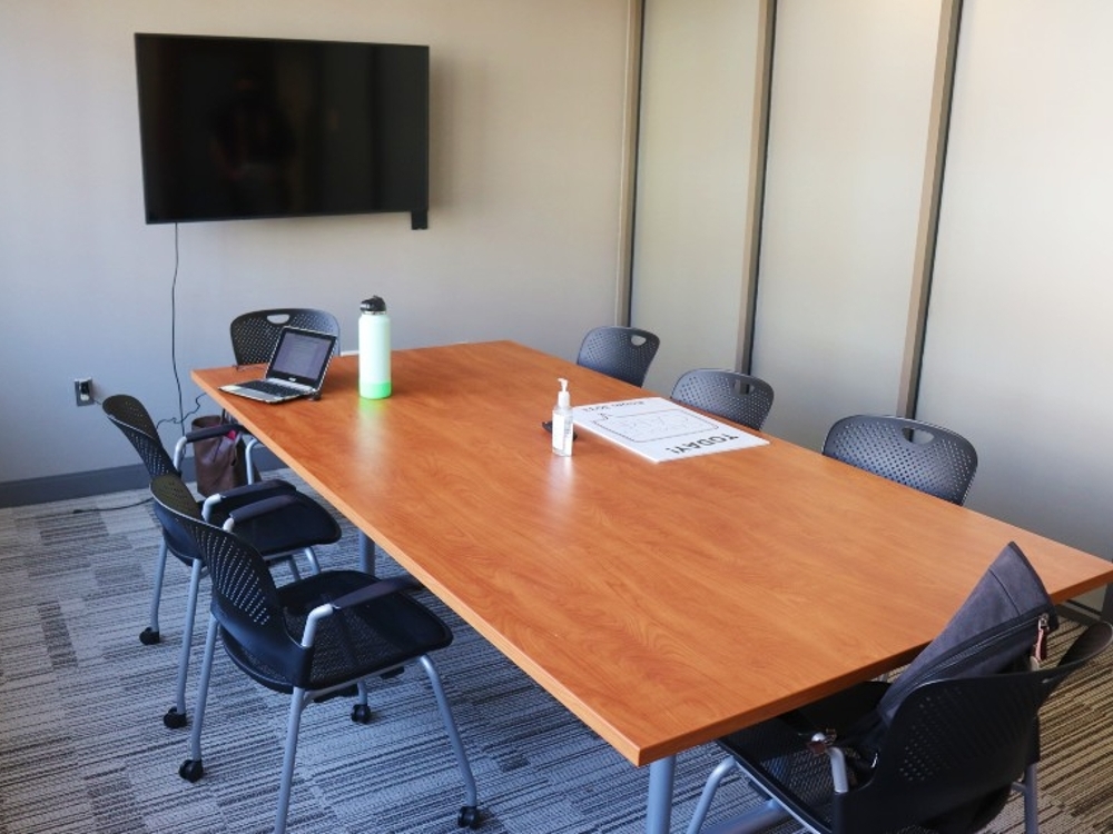 Meeting room with a flat screen tv on the wall, a large wooden table in the center, and 7 chairs around the able.