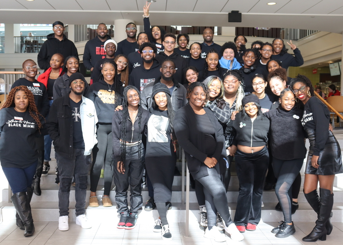 Large group of Black students wearing black clothing, smiling and posing on the steps of the Armstrong food court area. 