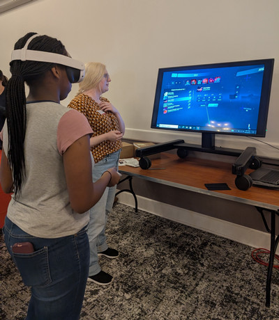 Two people test a virtual reality setup, with one wearing a VR headset and a monitor showing the VR interface on a table.