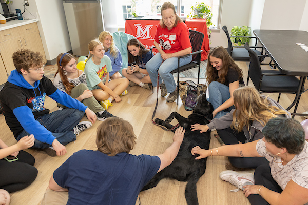 Group of students sitting in a circle and petting a black therapy dog during a school visit.