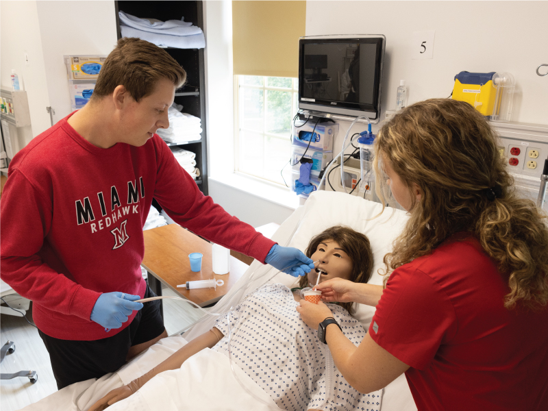 Two nursing students standing beside a hospital bed practicing patient care on a nursing mannequin