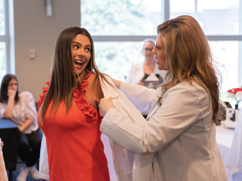 Graduate nursing student putting on their white coat with the help of their professor.