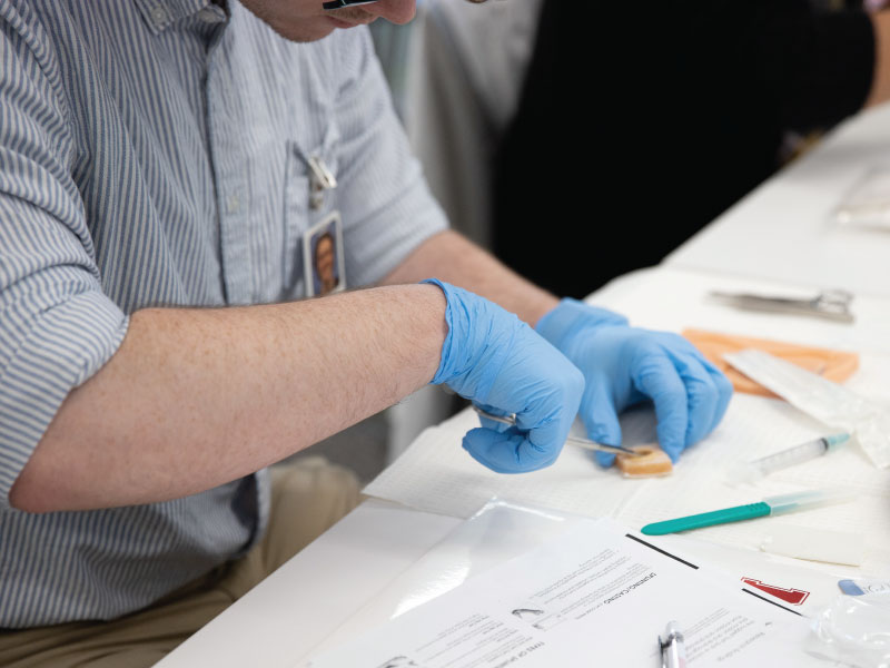 A MSN student with blue gloves on practicing how to insert a needle into a rubber body part during a skills workshop.