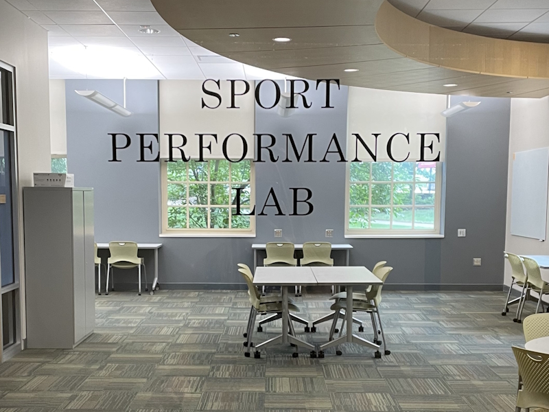 View of Sport Performance Lab at Miami University  with "Sport Performance Lab written across the image that includes a carpeted room with a file cabinet, desks and chairs and two windows with lovely views of the campus..