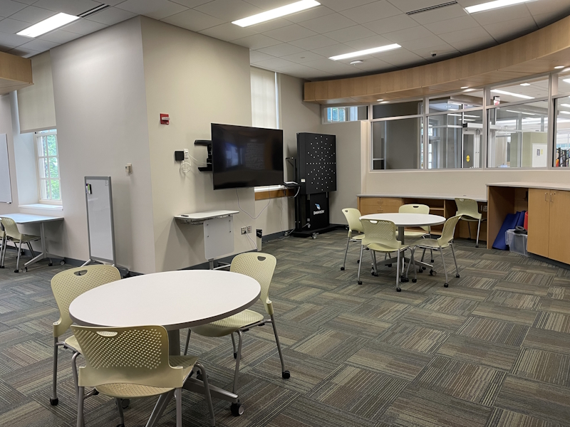 View of Sport Performance Lab at Miami University, a carpeted room with storage cabinets, desks and chairs, whiteboard, wall-mounted video screen, lab equipment and a wall of windows looking out to the Laws Hall's building atrium.