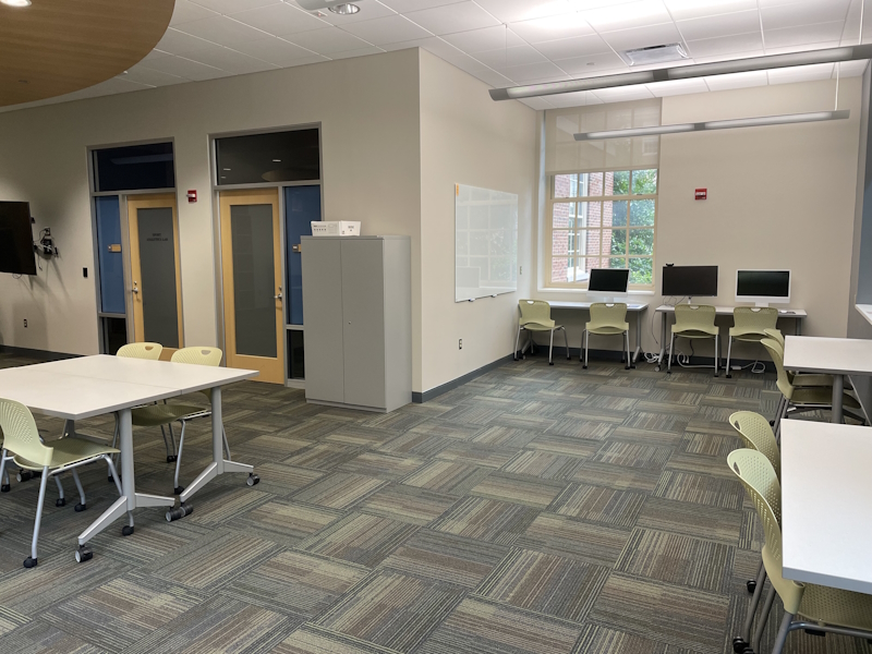 View of Sport Performance Lab at Miami University that includes a carpeted room with a file cabinet, desks and chairs, computer workstations, a wall-mounted video screen, two doors to two interior lab spaces and a window with views of the campus.