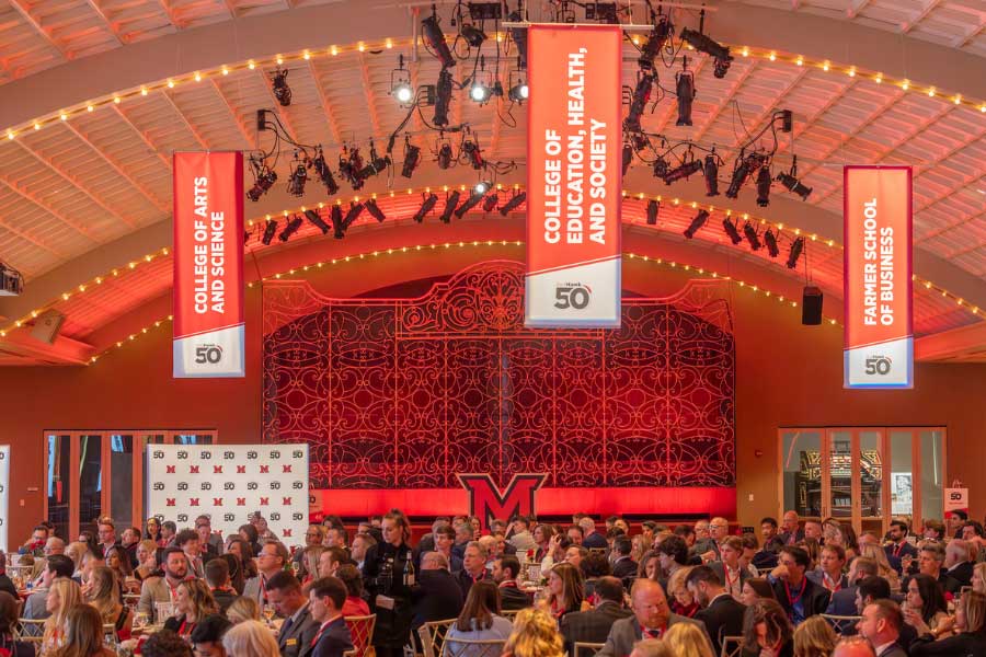 a large audience seated at round tables at a banquet hall in Music Hall with a streamer with the redHawk50 logo each for the College of Arts and Science, the College of Education, Health, and Society, and Farmer School of Business above the attendees
