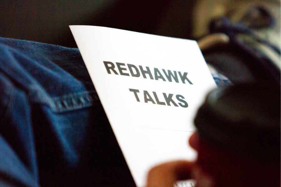 a paper with the words Redhawk Talks balanced on a persons crossed leg with their coffee mug out of focus
