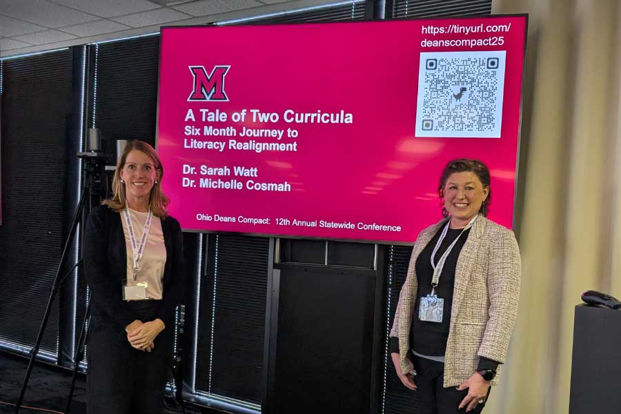 Sarah Watt and Michelle Cosmah posing on the sides of a presentation screen displaying the title slide of their presentation