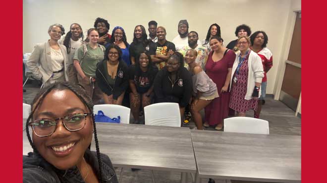 A group of about 18 smiling people, including students from the TEACh Cincinnati program and representatives from Cincinnati Public Schools’ Human Resources team, pose together in a classroom setting. Sydnie Singleton, a Miami University staff member, is in the foreground is taking a selfie with the group in the background. The room has neutral-colored walls, a whiteboard behind them, and gray tables and chairs in front. Most people are casually dressed, and the atmosphere appears cheerful and welcoming.