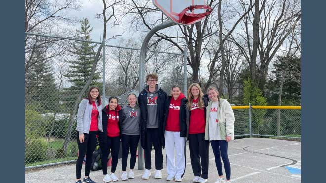 Seven college students stand together beneath a basketball hoop on an outdoor court, smiling in front of a chain-link fence and trees. Most are wearing Miami University apparel. The group includes participants and volunteers from the “Recess for Champions” program at Kramer Elementary, which promotes life skill development through sport-based play.