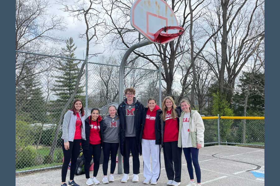 Seven college students stand together beneath a basketball hoop on an outdoor court, smiling in front of a chain-link fence and trees. Most are wearing Miami University apparel. The group includes participants and volunteers from the “Recess for Champions” program at Kramer Elementary, which promotes life skill development through sport-based play.